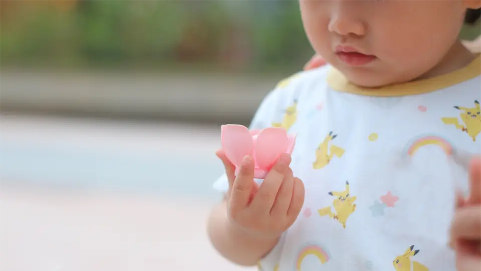 hanabii-pacifier-ergonomics-72431.webp طفل صغير يرتدي قميصًا يحمل صورة بيكاتشو ويحمل لهاية هانابي وردية اللون، ويفحص تصميمها على شكل بتلات بفضول.