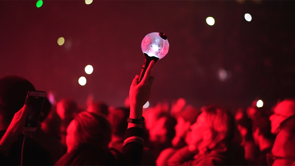 k-pop-lightstick-73531.webp Une personne brandissant un bâton lumineux de K-pop dans la foule d’un concert, entourée de fans illuminés par l’éclairage rouge de la scène et des points lumineux diffus en arrière-plan.
